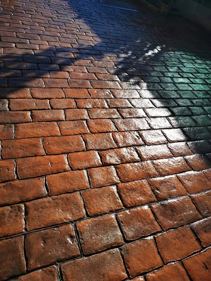 Pavimento de hormigón impreso con diseño que reproduce la apariencia del adoquín tradicional, con acabado en tonos rojizos y textura marcada para exteriores.