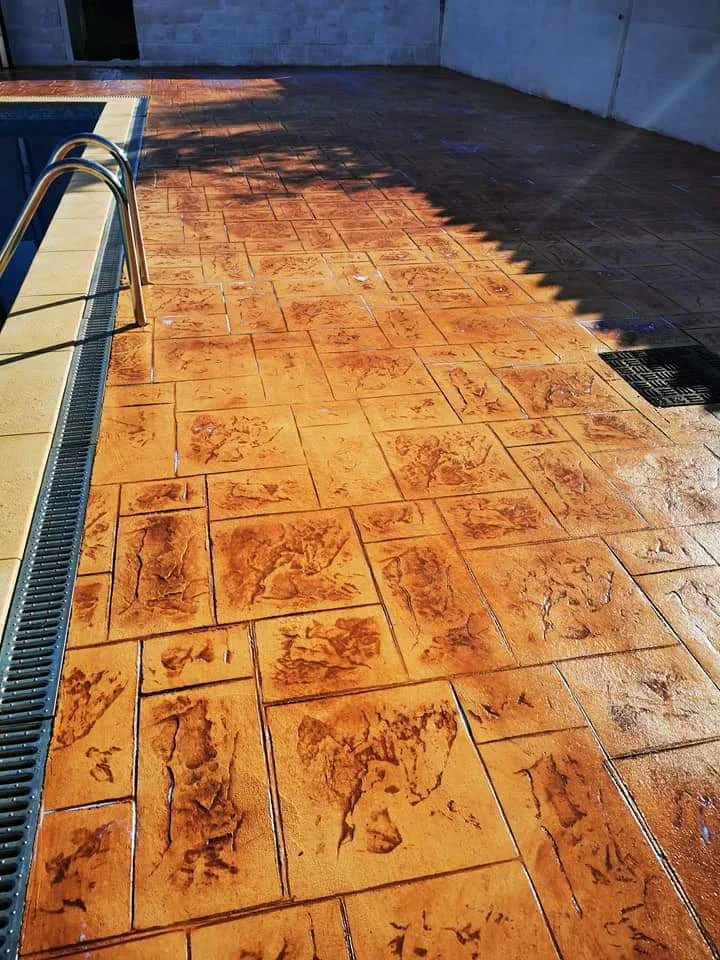 Pavimento exterior de hormigón impreso con acabado decorativo en tonos cálidos, aplicado en la zona de piscina para conseguir una superficie resistente, duradera y de fácil manteni