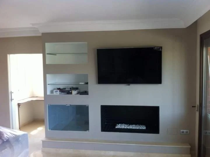 Resultado final de un mueble de pladur a medida, completamente acabado y pintado, con televisión suspendida, chimenea integrada y estanterías decorativas.