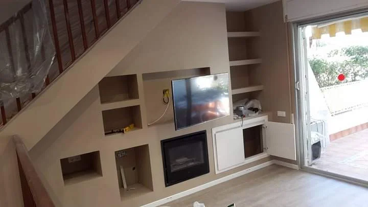 Resultado final de un mueble de pladur completamente integrado bajo la escalera, con televisión, chimenea, estanterías y espacios de almacenamiento a medida.