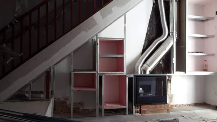 Construcción de una estructura metálica de pladur adaptada al hueco bajo la escalera, diseñada para integrar almacenamiento, estanterías, televisión y chimenea.